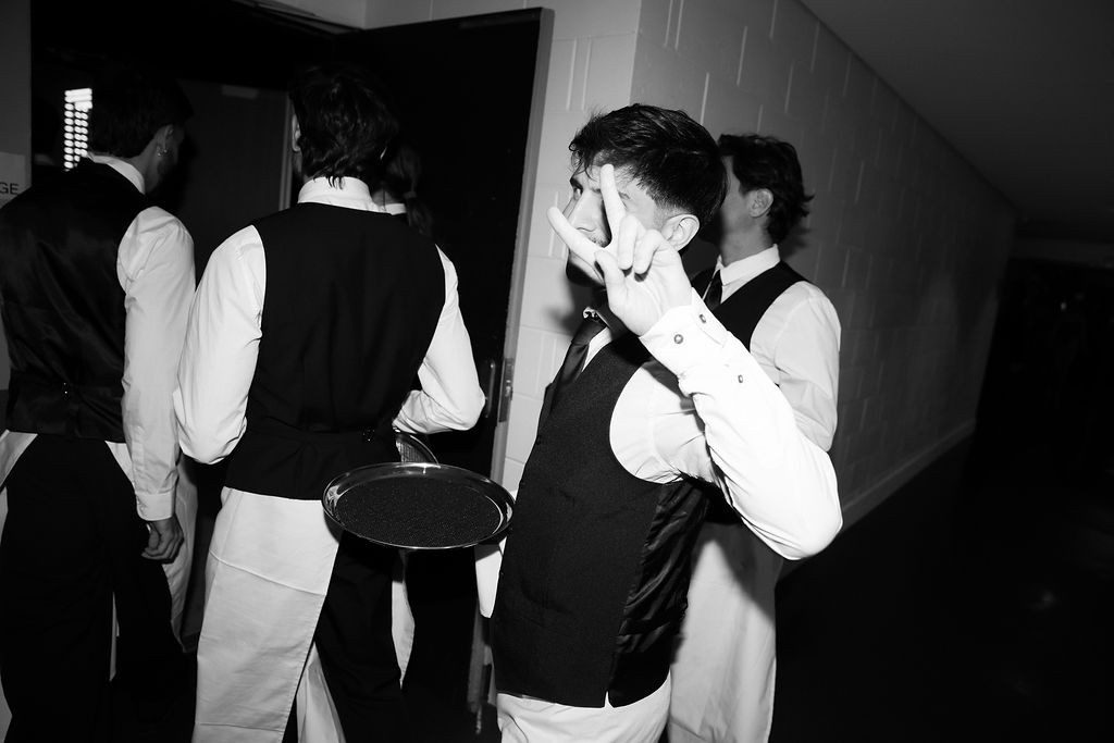 Waitstaff in black tie gathered before service, one giving a discreet signal